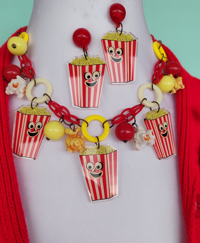 May include: A quirky jewellery set with a necklace and earrings. The necklace and earrings feature popcorn box charms with smiling faces, red and white stripes, and yellow popcorn. The necklace has red chain links, yellow and red beads.