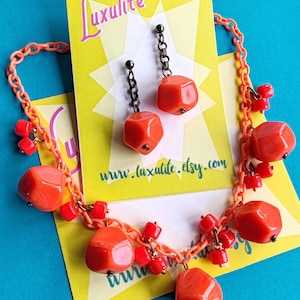 May include: A necklace and a pair of earrings featuring bright orange, faceted, irregular-shaped beads on a light orange plastic chain. The jewellery is displayed on yellow cards with the text Luxulite and www.luxulite.etsy.com printed in script.