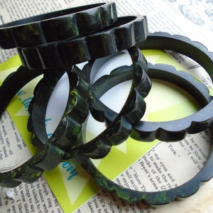 May include: A collection of dark green, scalloped-edge bangles. The bracelets have a marbled appearance with hints of yellow and are stacked on top of each other. The background includes vintage paper and a retro-style card.