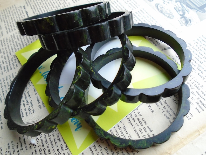 May include: A collection of dark green, scalloped-edge bangles. The bracelets have a marbled appearance with hints of yellow and are stacked on top of each other. The background includes vintage paper and a retro-style card.