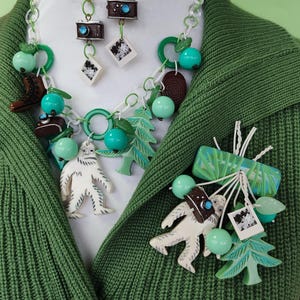 May include: A quirky necklace and earring set with a white chain, green details, and assorted charms. Charms include a camera, a yeti, a tree, and green beads. The set is shown on a green cardigan.
