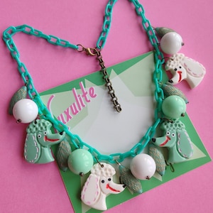 May include: A turquoise chain necklace featuring poodle charms, white and mint green beads, and leaf-shaped details. The poodle charms are white and mint green with red accents. The necklace is set against a pink backdrop.