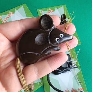 Könnte beinhalten: Ein dunkelbrauner mausförmiger Anhänger mit weißen Augen und goldenen Schnurrhaaren. Die Maus hat einen hellbraunen geflochtenen Schwanz. Der Anhänger wird in einer Hand gehalten, mit anderen ähnlichen Anhängern und Karten im Hintergrund.