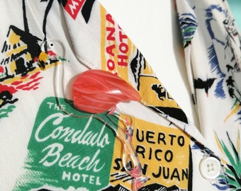 May include: A flamingo-shaped brooch with a red and clear design, pinned to a white shirt with a tropical print. The shirt features text and illustrations of hotels and destinations like Miami and Puerto Rico.