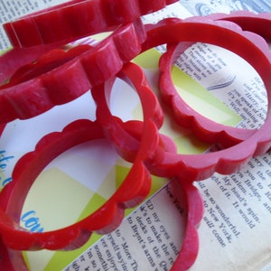 May include: Several red, scalloped-edge bangles are scattered across a vintage book page. The bright red color of the bracelets contrasts with the aged paper, creating a vintage aesthetic. The bangles are approximately 2.5 inches in diameter.
