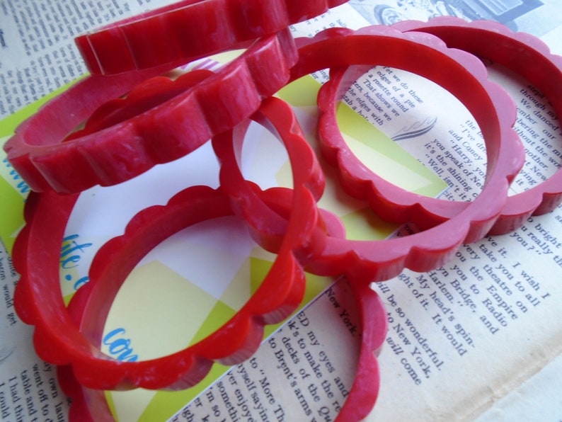 May include: Several red, scalloped-edge bangles are scattered across a vintage book page. The bright red color of the bracelets contrasts with the aged paper, creating a vintage aesthetic. The bangles are approximately 2.5 inches in diameter.