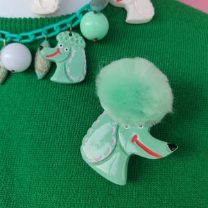 Puede incluir: Un broche de caniche verde con un pomp&oacute;n esponjoso y un collar a juego. El collar presenta dijes de caniche, cuentas y una cadena verde. Los dijes de caniche son verdes y blancos con detalles en rosa.