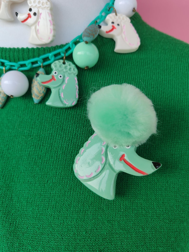 Puede incluir: Un broche de caniche verde con un pomp&oacute;n esponjoso y un collar a juego. El collar presenta dijes de caniche, cuentas y una cadena verde. Los dijes de caniche son verdes y blancos con detalles en rosa.