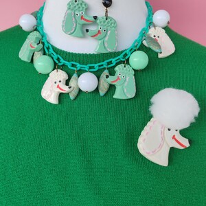 May include: A vintage-style jewelry set featuring a poodle theme. The set includes a necklace, earrings, and a brooch. The necklace and earrings have green poodle pendants, while the brooch has a white poodle. The necklace has white and green beads.