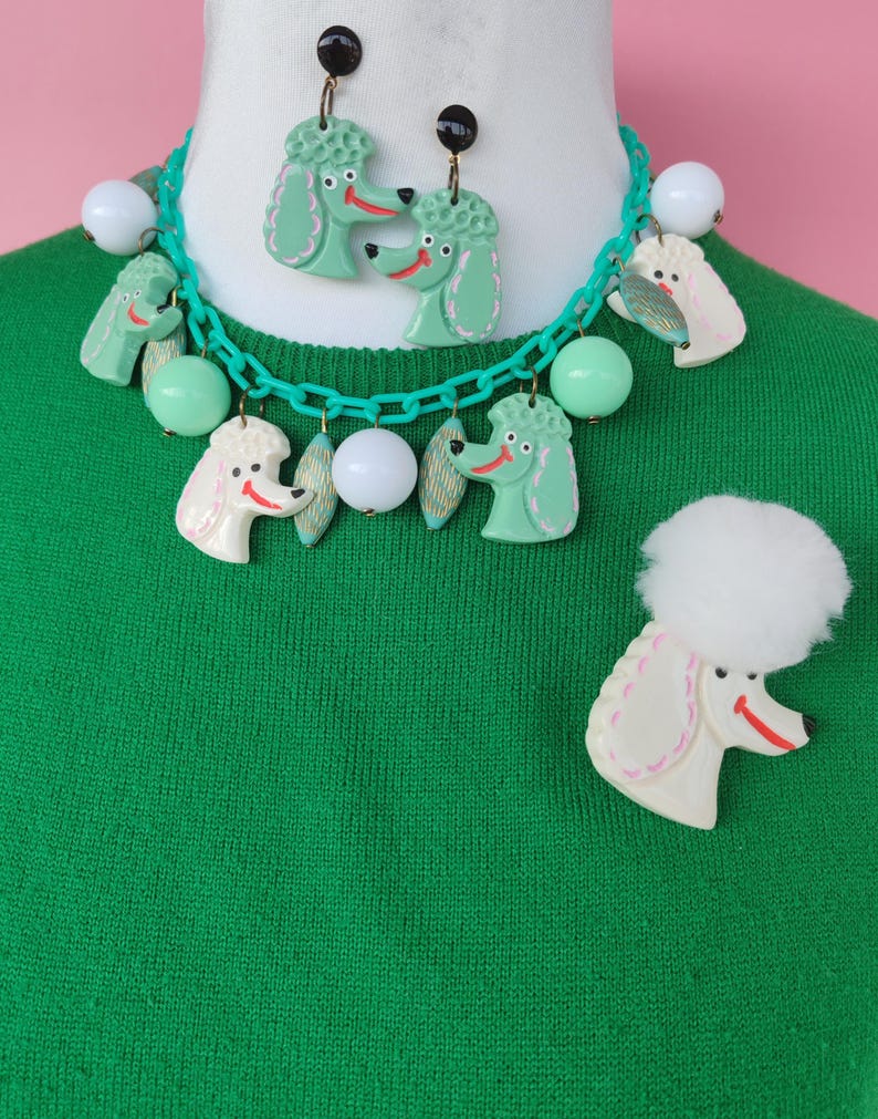 May include: A vintage-style jewelry set featuring a poodle theme. The set includes a necklace, earrings, and a brooch. The necklace and earrings have green poodle pendants, while the brooch has a white poodle. The necklace has white and green beads.