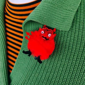 May include: A red devil-shaped brooch with black details is pinned to a green knitted cardigan. The brooch has a fluffy texture and a cartoon design. Beneath, a black and orange striped top is visible.