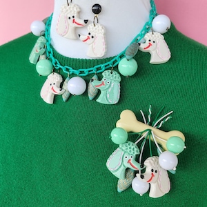 May include: A vintage-style jewelry set featuring poodle designs. The set includes a necklace, earrings, and a brooch. The necklace and earrings have white poodle heads with red smiles and black accents. The brooch has a bone and poodle design.