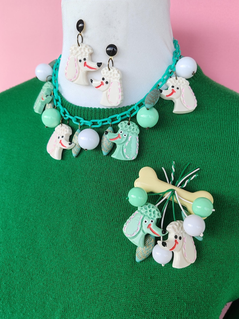 May include: A vintage-style jewelry set featuring poodle designs. The set includes a necklace, earrings, and a brooch. The necklace and earrings have white poodle heads with red smiles and black accents. The brooch has a bone and poodle design.