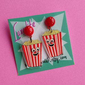May include: Novelty earrings with a popcorn design. Each earring has a red and white striped popcorn container with a smiling face and yellow popcorn. The earrings are attached to a red ball stud. The earrings are on a teal card with the text "Luxelite" and "kulite.etsy.com".
