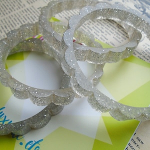 May include: Four clear, glitter-infused bangles with a scalloped edge. The bracelets are transparent with silver glitter, creating a sparkling effect. The bangles are stacked on top of each other, showcasing their design.