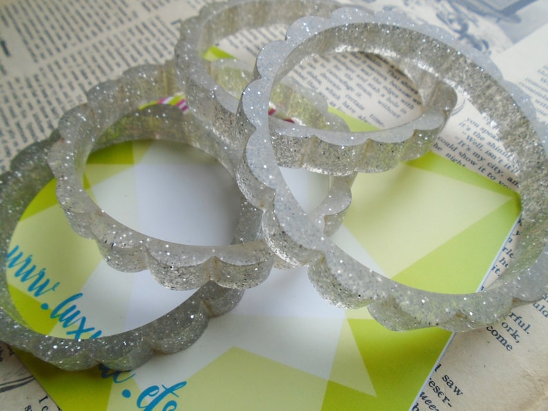 May include: Four clear, glitter-infused bangles with a scalloped edge. The bracelets are transparent with silver glitter, creating a sparkling effect. The bangles are stacked on top of each other, showcasing their design.