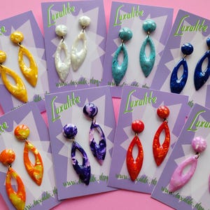 May include: A selection of colourful dangle earrings on purple cards. Each pair has a round stud and a long, open teardrop shape. Colours include yellow, white, turquoise, blue, orange, purple, red, and pink. The cards have the text "Luxulite".