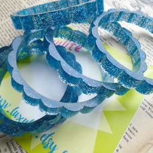 May include: Several blue glitter bangles with a scalloped edge are stacked on top of each other. The bangles are made of a translucent material and are scattered on a background of vintage paper and a green and white card.