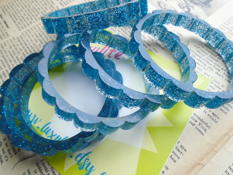 May include: Several blue glitter bangles with a scalloped edge are stacked on top of each other. The bangles are made of a translucent material and are scattered on a background of vintage paper and a green and white card.