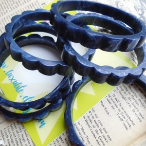 May include: Several dark blue bangles with a scalloped edge are stacked on a vintage newspaper background. The bangles have a marbled appearance and are made of a solid material. The website address "www.luxulite.etsy.com" is visible on a yellow card.