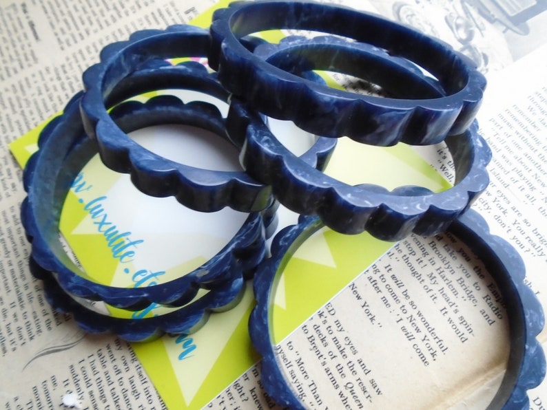 May include: Several dark blue bangles with a scalloped edge are stacked on a vintage newspaper background. The bangles have a marbled appearance and are made of a solid material. The website address "www.luxulite.etsy.com" is visible on a yellow card.