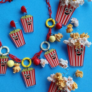May include: A quirky jewellery set comprising a red chain necklace and earrings. The necklace and earrings feature popcorn bucket charms with smiling faces, red and white stripes, and yellow popcorn details. The background is a solid blue.