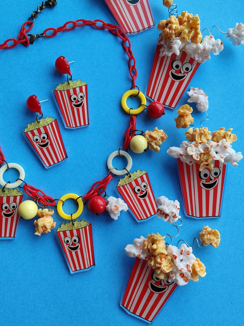 May include: A quirky jewellery set comprising a red chain necklace and earrings. The necklace and earrings feature popcorn bucket charms with smiling faces, red and white stripes, and yellow popcorn details. The background is a solid blue.