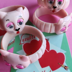 May include: Four vintage pink Luxulite cat face bangles. Each bangle features a cat head with pink eyeshadow, black whiskers, and a red mouth. The bangles are on a pink background with a green card that has two red hearts and the website address.