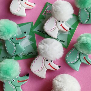 Puede incluir: Broches con forma de caniche, en blanco y verde menta, cada uno con un pomp&oacute;n esponjoso y una cara sonriente. Los broches est&aacute;n sobre un fondo rosa, con tarjetas de visita que dicen "www.luxulite.etsy.com".