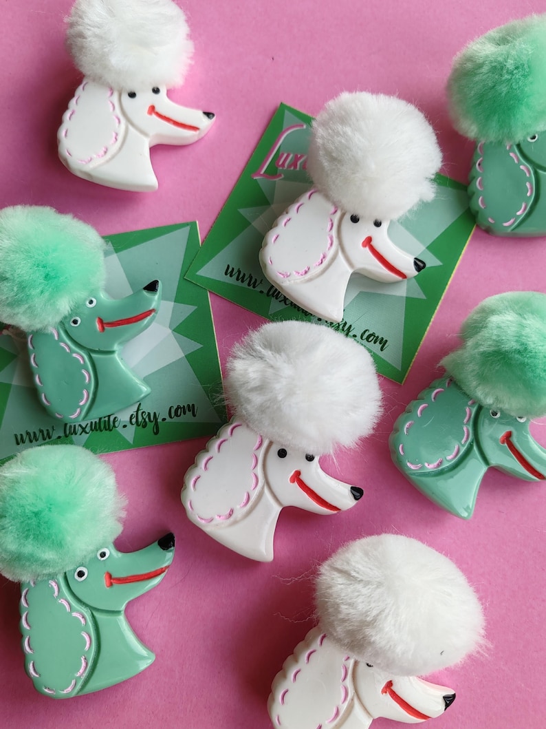 Puede incluir: Broches con forma de caniche, en blanco y verde menta, cada uno con un pomp&oacute;n esponjoso y una cara sonriente. Los broches est&aacute;n sobre un fondo rosa, con tarjetas de visita que dicen "www.luxulite.etsy.com".