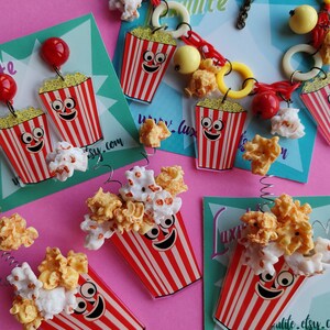 May include: Novelty jewellery and accessories with a popcorn theme. The items include earrings, a necklace, and charms, all with a red and white striped popcorn bucket design, filled with popcorn.