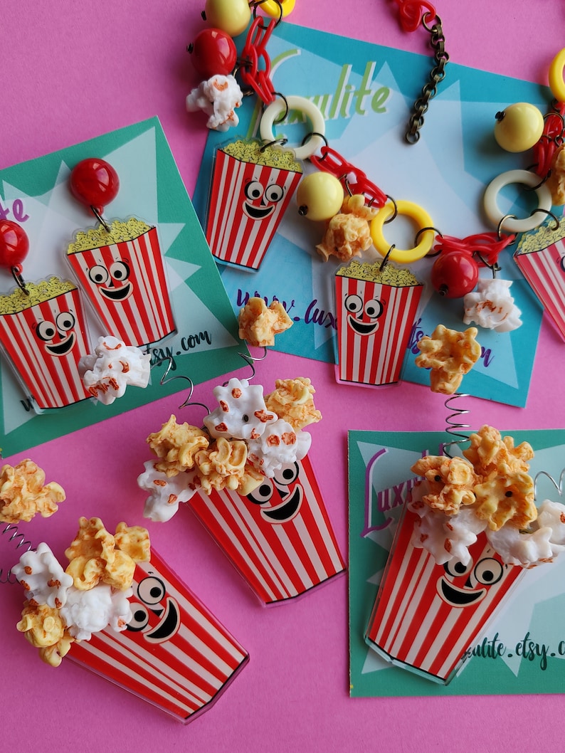 May include: Novelty jewellery and accessories with a popcorn theme. The items include earrings, a necklace, and charms, all with a red and white striped popcorn bucket design, filled with popcorn.