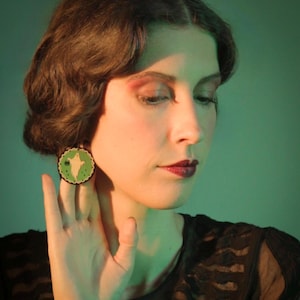 Op de afbeelding: Een vrouw met donker haar draagt een zwarte top en een ronde oorbel met een groen en wit spookontwerp. De oorbel is een Halloween-accessoire. De vrouw raakt haar nek aan met haar hand.