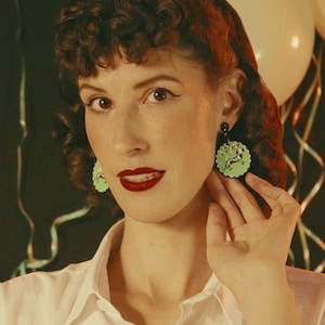 May include: Round dangle earrings featuring a light green scalloped edge and a central illustration of a brown and white dachshund dog. The earrings are suspended from a small black circular stud component, creating a distinct contrast in color.