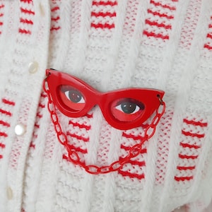 Pode incluir: Broche de plástico vermelho em forma de óculos de gato com corrente, preso a um cardigã de malha branco e vermelho estampado. Os óculos têm olhos pintados. O cardigã tem um design de malha texturizada com listras vermelhas.
