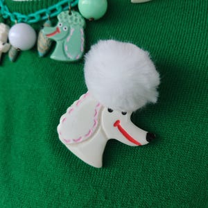 Puede incluir: Un broche de caniche blanco con un pomp&oacute;n blanco esponjoso, una sonrisa roja y una nariz negra. El broche est&aacute; sobre un fondo verde, con un collar a juego con dijes de caniche y cuentas verdes.