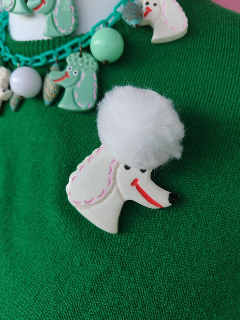 Puede incluir: Un broche de caniche blanco con un pomp&oacute;n blanco esponjoso, una sonrisa roja y una nariz negra. El broche est&aacute; sobre un fondo verde, con un collar a juego con dijes de caniche y cuentas verdes.