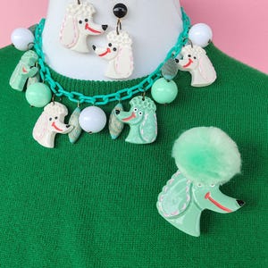 Puede incluir: Un conjunto de joyas de estilo vintage con dise&ntilde;os de caniches. El conjunto incluye un collar con eslabones de cadena verdes y dijes de caniche, aretes a juego y un broche de caniche. Los caniches son blancos y verde menta con detalles rojos.