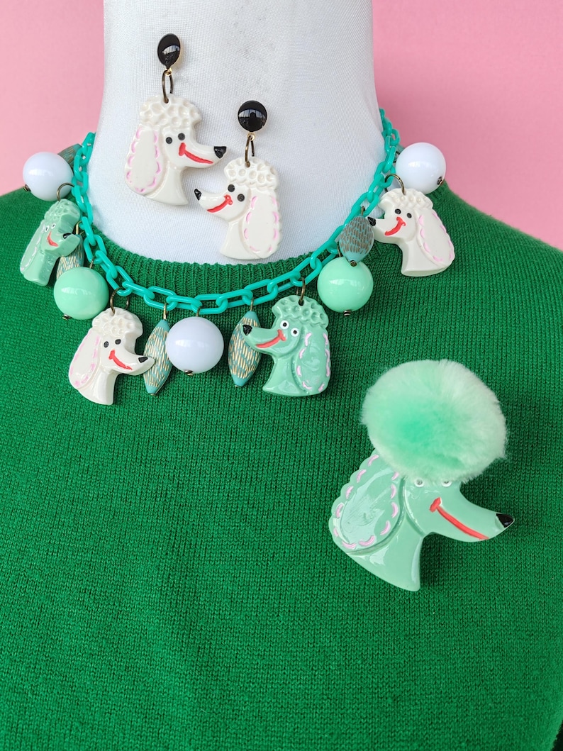 Puede incluir: Un conjunto de joyas de estilo vintage con dise&ntilde;os de caniches. El conjunto incluye un collar con eslabones de cadena verdes y dijes de caniche, aretes a juego y un broche de caniche. Los caniches son blancos y verde menta con detalles rojos.