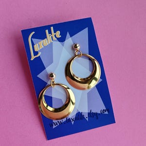 May include: Gold-coloured, half-circle hoop earrings with a glossy finish. The earrings are presented on a blue card with the text "Luxalite" and a website address. The background is a solid pink colour.