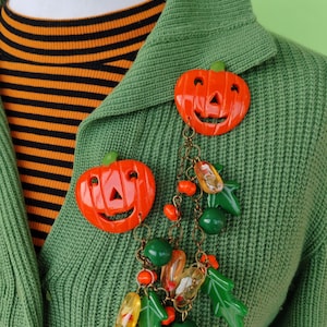 Op de afbeelding: Twee oranje pompoenvormige broches met groene stelen en lachende gezichten. Ze zijn verbonden door een ketting met groene bladeren, oranje kralen en heldere, gele en oranje druppelvormige bedels. De broches zijn vastgemaakt aan een groene trui.