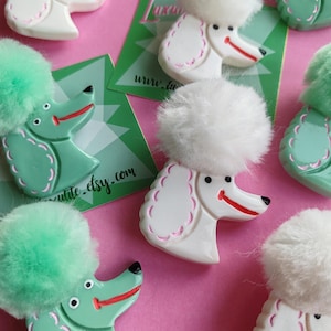 Puede incluir: Broches con forma de caniche en blanco y verde menta, cada uno con un pomp&oacute;n esponjoso y una sonrisa roja. Los broches est&aacute;n dispuestos sobre un fondo rosa, con tarjetas de visita.