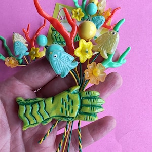 Puede incluir: Un ramo decorativo que presenta una mano de criatura verde, ramas de coral naranja, flores amarillas y naranjas, dijes en forma de pez azul claro y amarillo, y cuentas redondas sobre tallos de alambre trenzado verde y amarillo.
