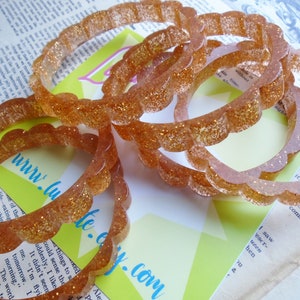 May include: Several clear, glitter-infused bangles with a scalloped edge are displayed. The bracelets are a warm, amber color and catch the light, creating a sparkling effect. The background includes text and a website address.