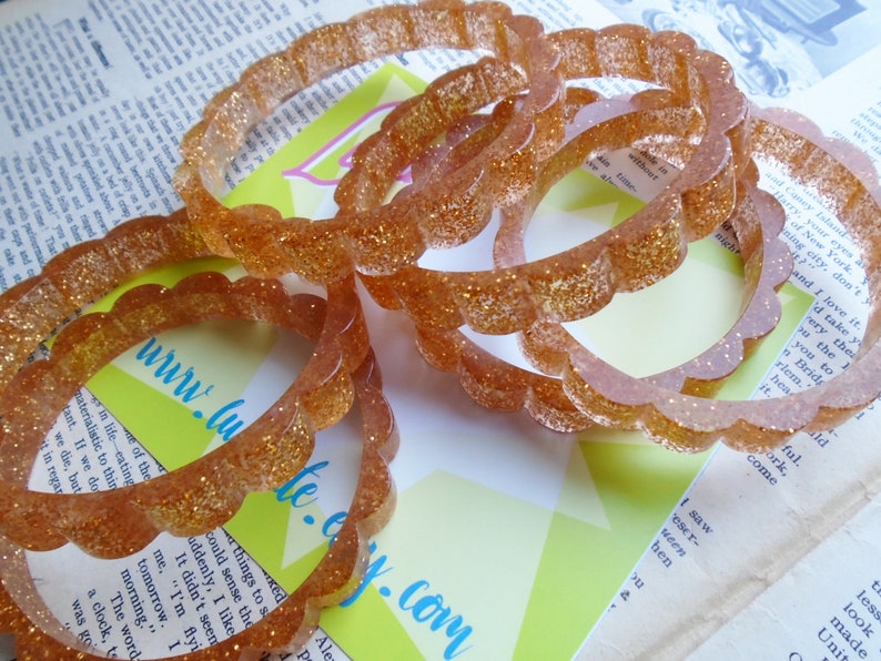 May include: Several clear, glitter-infused bangles with a scalloped edge are displayed. The bracelets are a warm, amber color and catch the light, creating a sparkling effect. The background includes text and a website address.