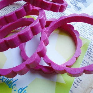 May include: Several bright pink bangles with a scalloped edge are stacked on top of each other. The bracelets are made of a solid material and are set against a backdrop of vintage paper and a yellow and green starburst design.