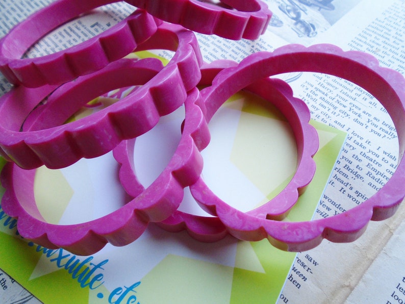 May include: Several bright pink bangles with a scalloped edge are stacked on top of each other. The bracelets are made of a solid material and are set against a backdrop of vintage paper and a yellow and green starburst design.