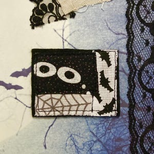 May include: A rectangular black and white Halloween-themed patch with the word "BOO" in white. The patch features a glittery black background, bat silhouettes, and a spiderweb design. Black lace and fabric scraps are in the background.