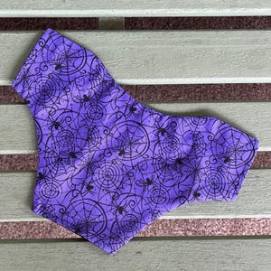 May include: A purple fabric accessory featuring a repeating black graphic print of spider webs and small spiders. The material has a distinct pattern of circular web designs interconnected with various arachnids across the entire surface area.