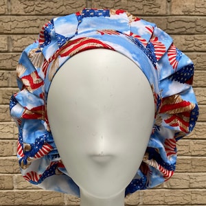 May include: A patriotic bouffant scrub cap featuring a repeating pattern of the American flag, eagles, and clouds. The cap is primarily blue, red, and white, with a gathered design for a comfortable fit. The cap is designed to cover the hair.